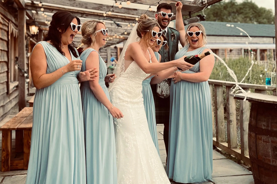 Bride, groom and bridesmaids wearing love heart shaped sunglasses pop bottle of champagne